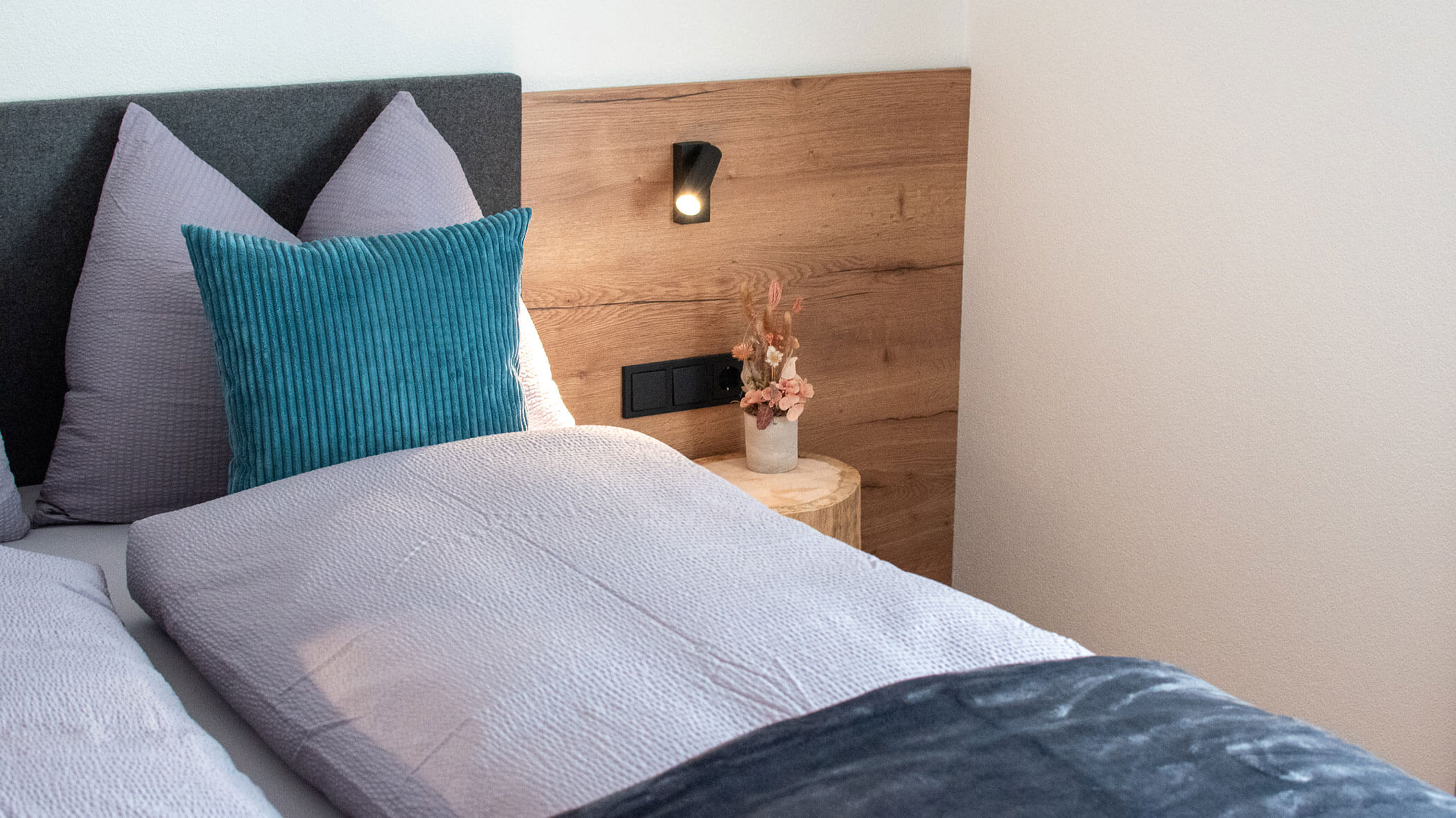 A neatly made bed with lavender bedding, a teal pillow, and a bedside table with a small vase of flowers. - Kerschbaumerhof