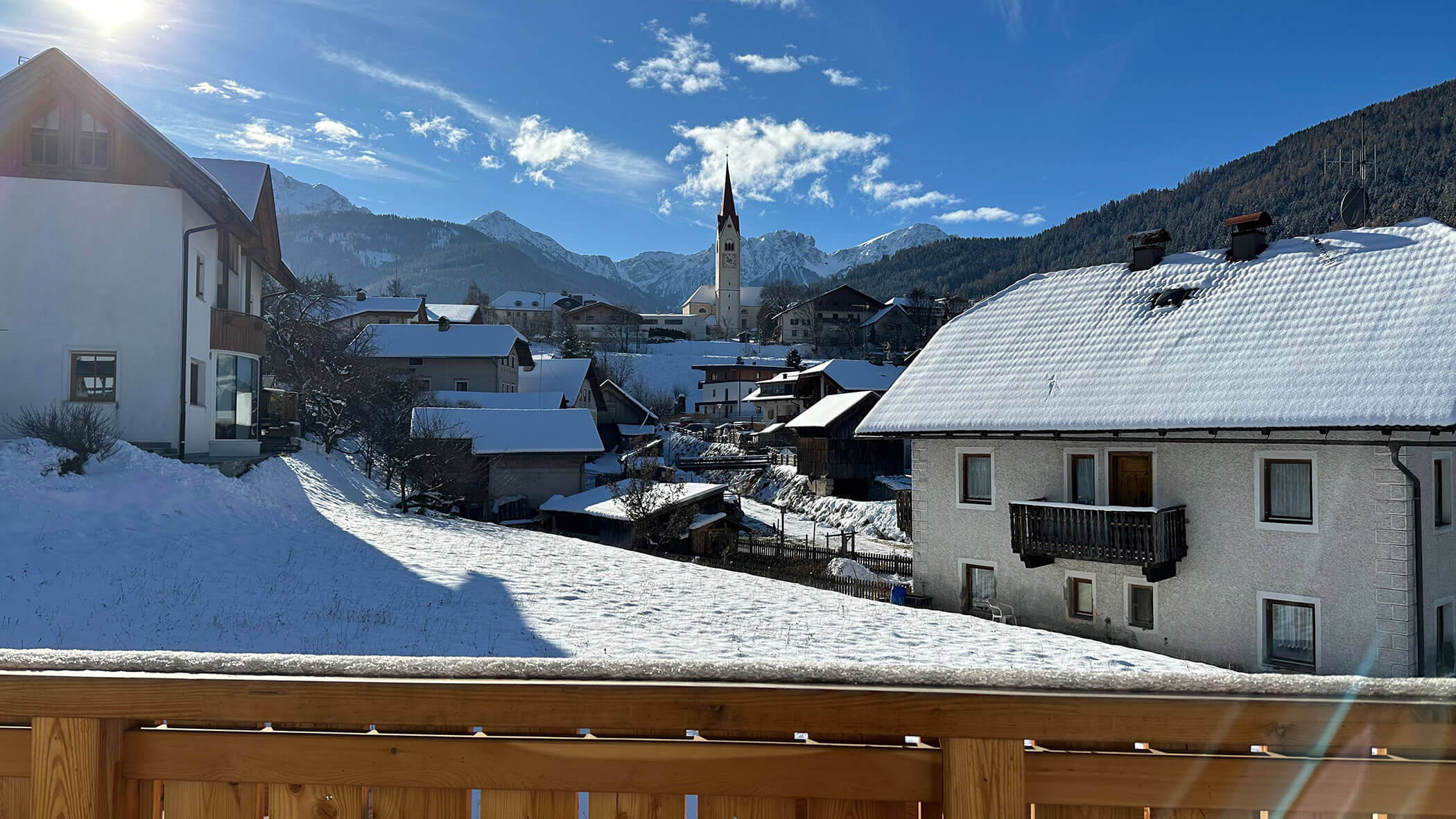 Snow-covered village with mountain backdrop, church tower, wooden fence, and clear blue sky. - Kerschbaumerhof