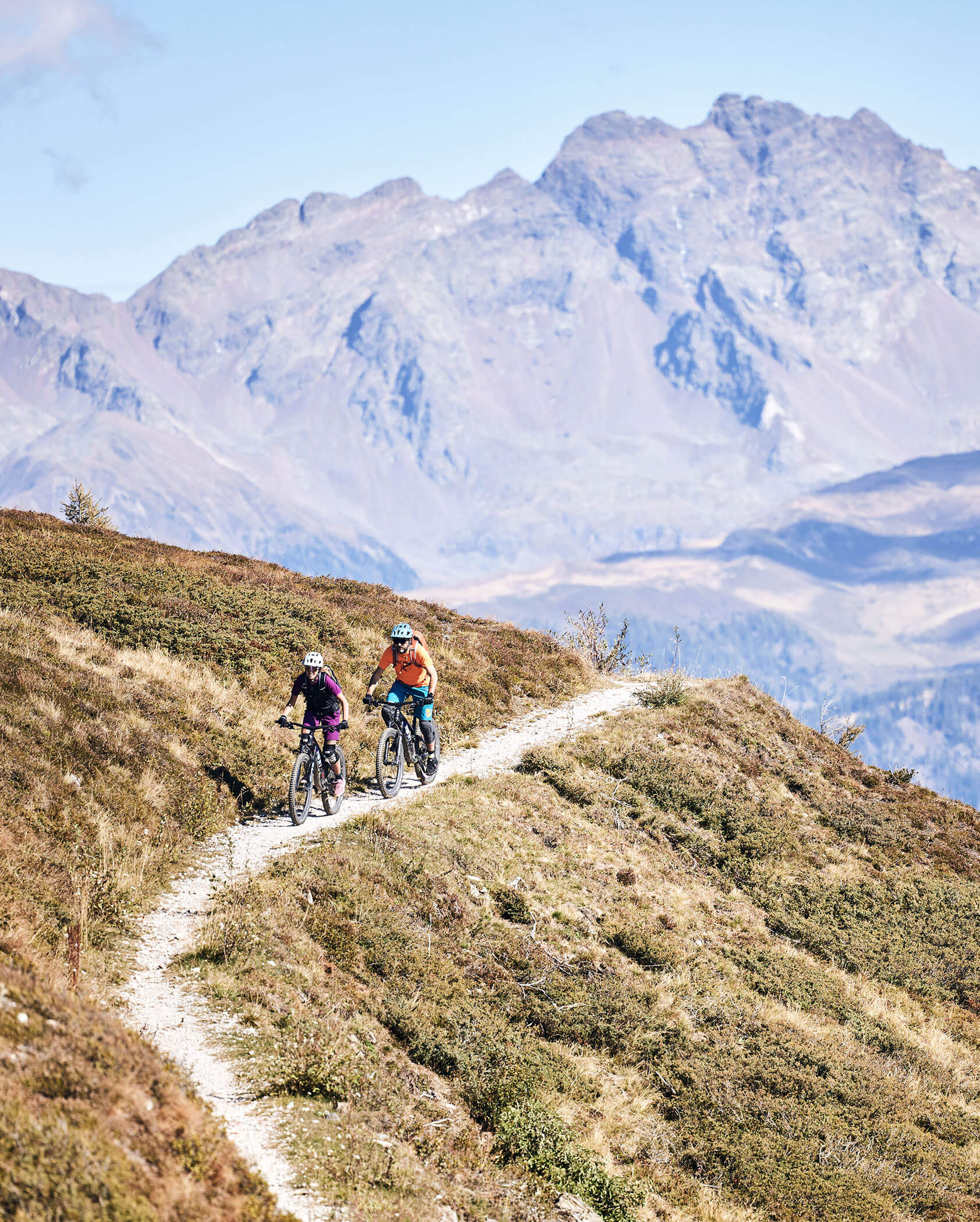 Zwei Personen fahren mit dem Mountainbike auf einem schmalen Weg mit Bergen im Hintergrund. - Kerschbaumerhof
