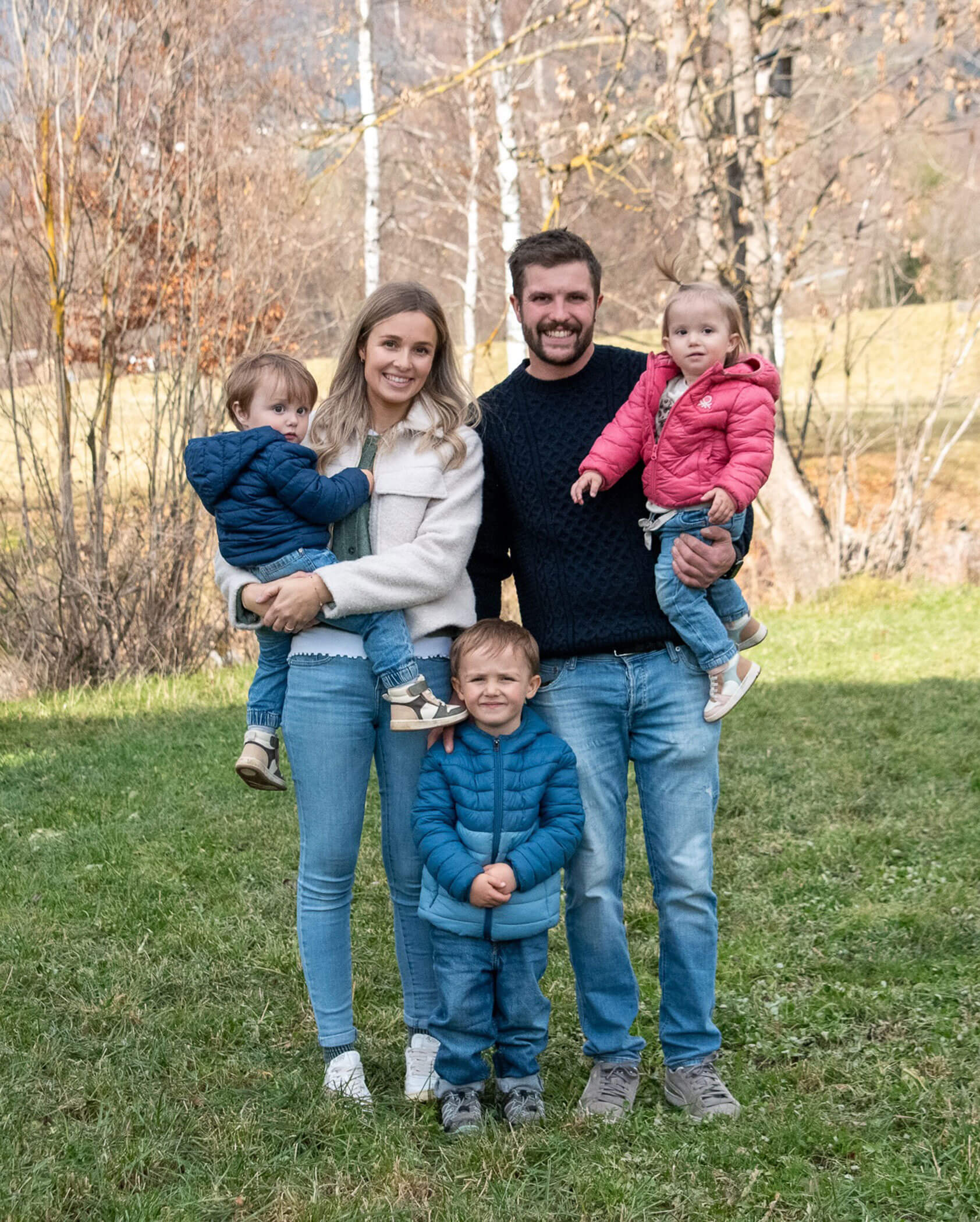 A family of five poses outside on grass, with parents holding two toddlers and an older child standing in front. - Kerschbaumerhof
