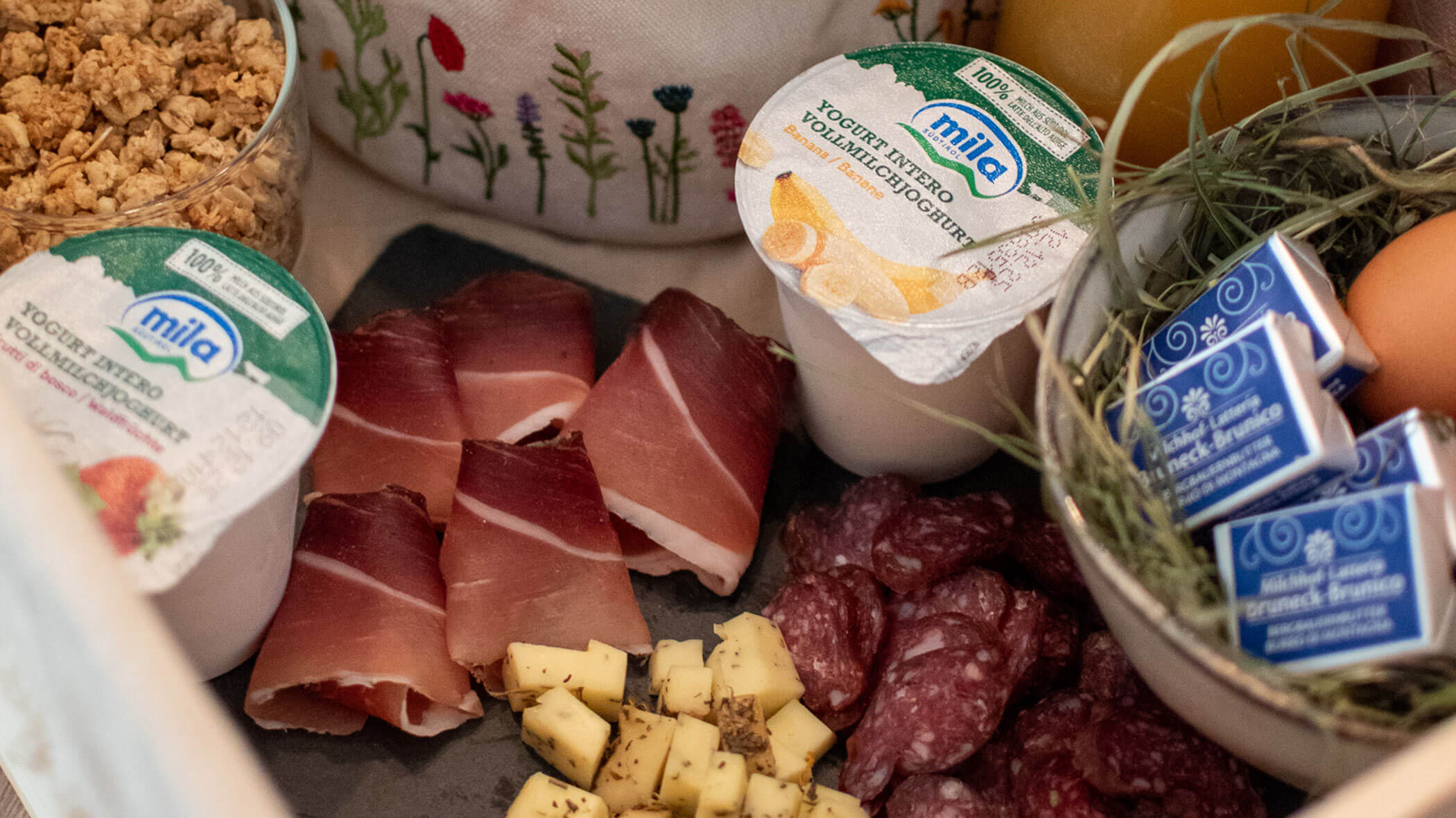 Ein Frühstückstablett mit Joghurt, Müsli, Wurstwaren, Käsewürfeln, Eiern und kleinen Kartons mit Sahne. - Kerschbaumerhof