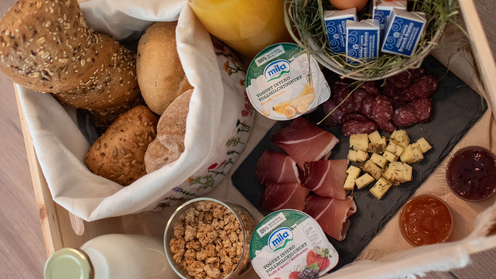Ein Holztablett mit Brot, Joghurt, Käse, Aufschnitt, Müsli, Marmelade, Butter, Eiern und Orangensaft. - Kerschbaumerhof