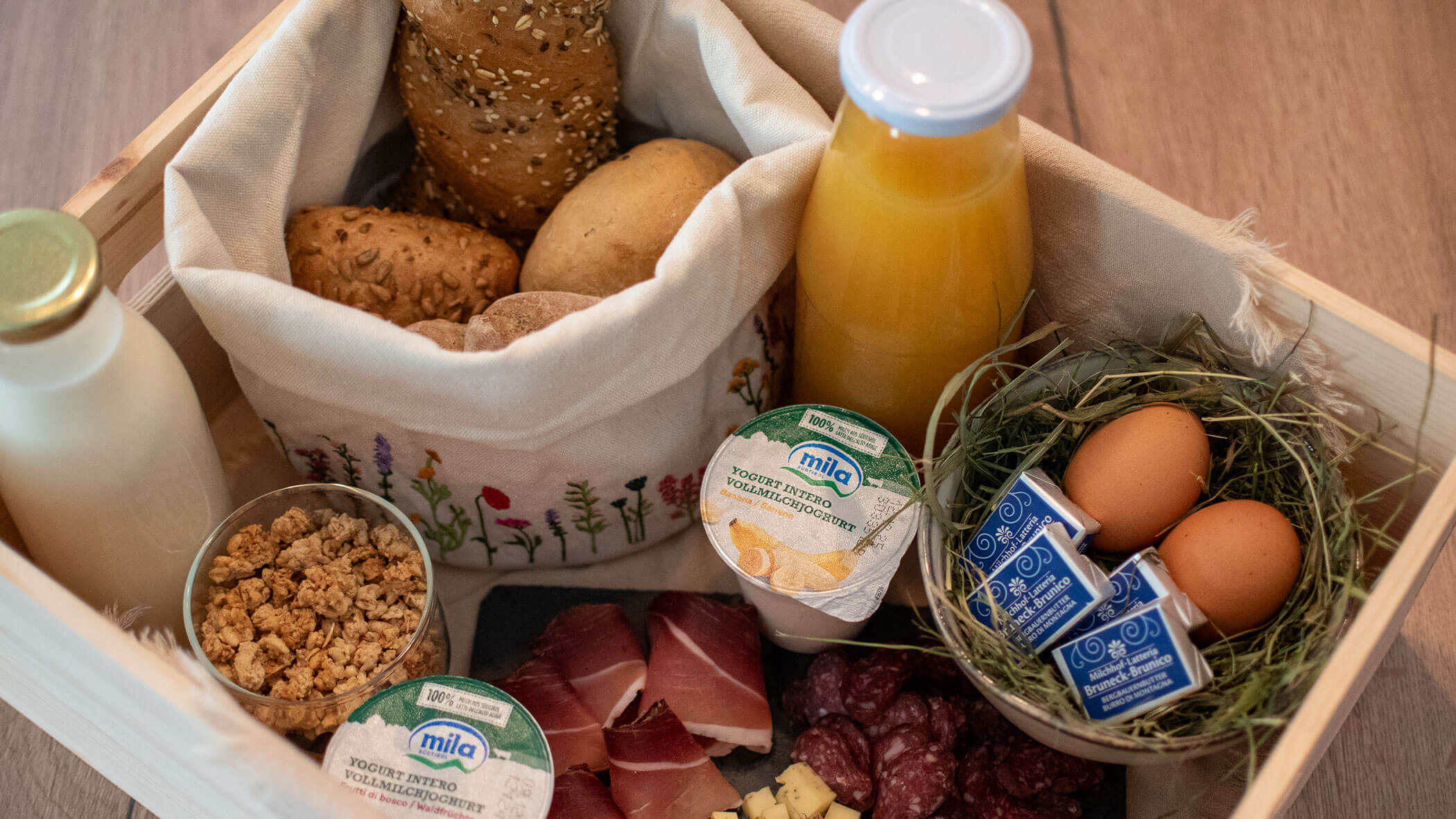 Frühstücksbox mit Brötchen, Eiern, Joghurt, Saft, Milch, Müsli, Käse und Wurstwaren in einer Holzkiste. - Kerschbaumerhof