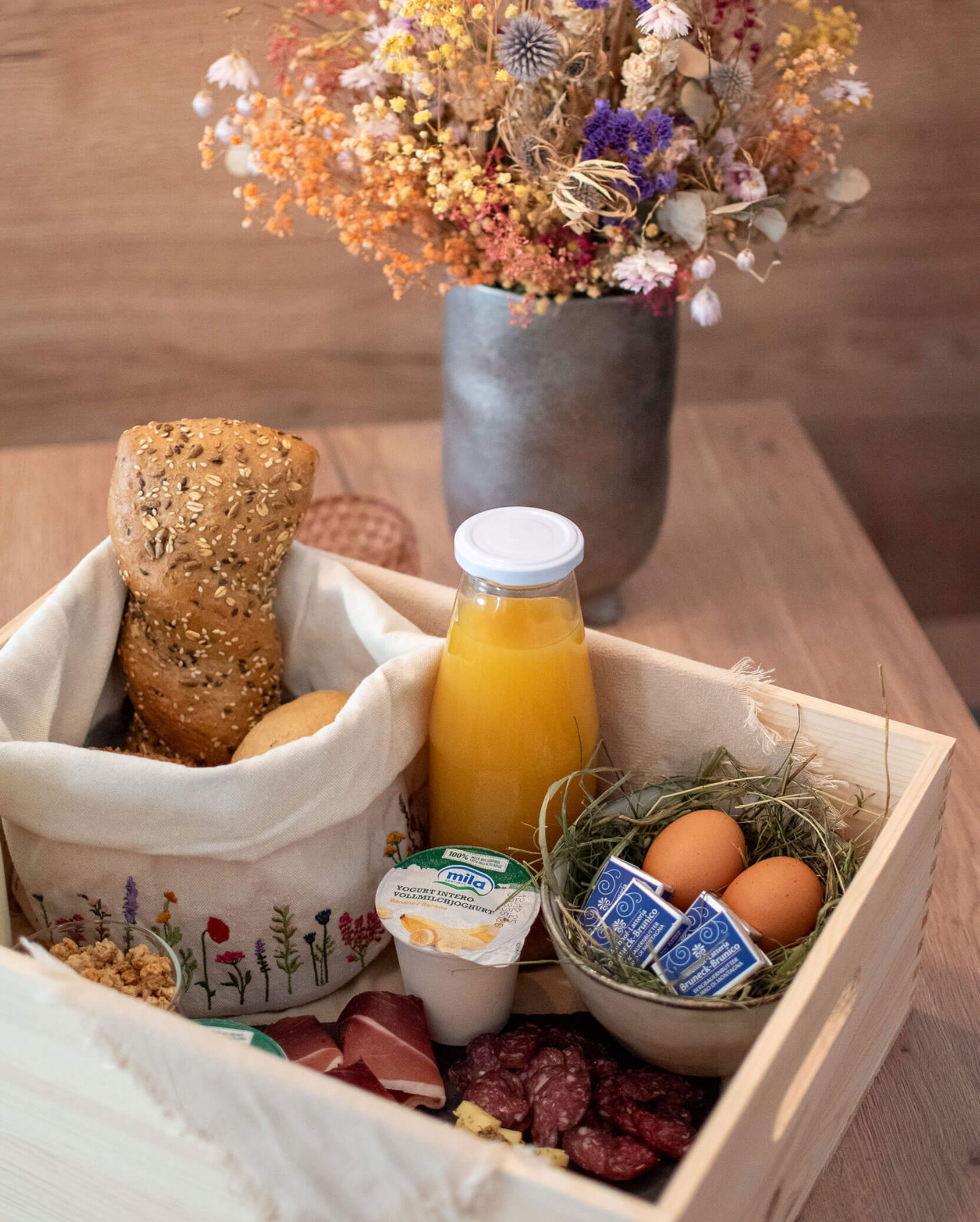 Breakfast box with bread, orange juice, eggs, yogurt, cold cuts, and flowers in a vase in the background. - Kerschbaumerhof