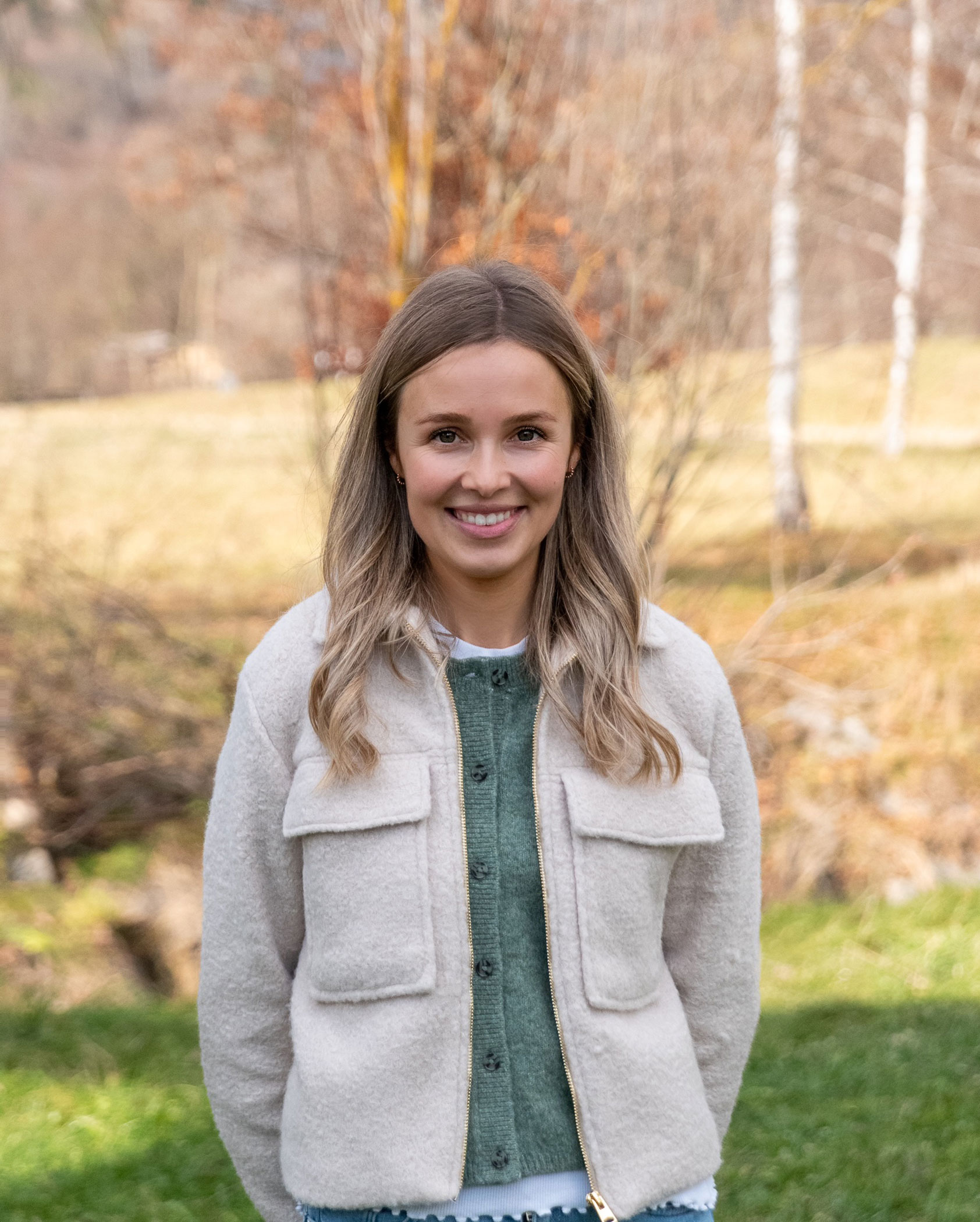 Eine junge Frau mit langen blonden Haaren lächelt im Freien auf einer Wiese mit Bäumen im Hintergrund. - Kerschbaumerhof