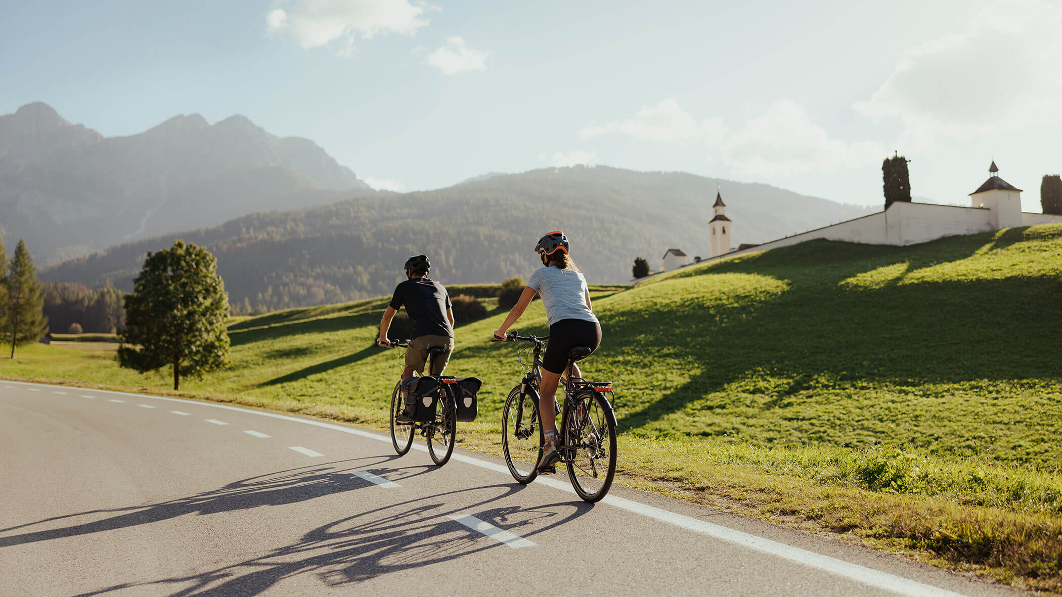 Two people ride bicycles on a scenic road, with green hills and mountains in the background. - Kerschbaumerhof