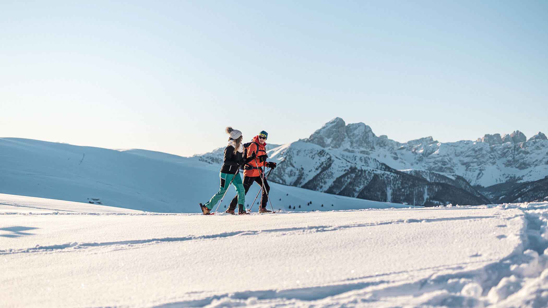Two people walk on snow with ski poles, surrounded by snowy mountains under a clear sky. - Kerschbaumerhof