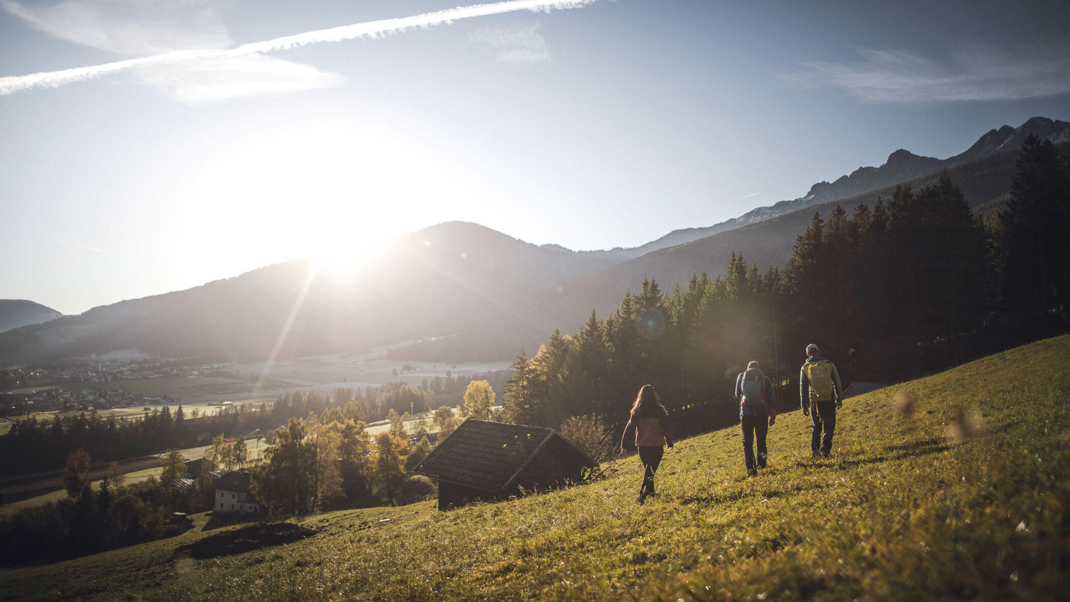 Three people hike uphill through a grassy meadow toward the sunrise, with mountains and trees in the background. - Kerschbaumerhof
