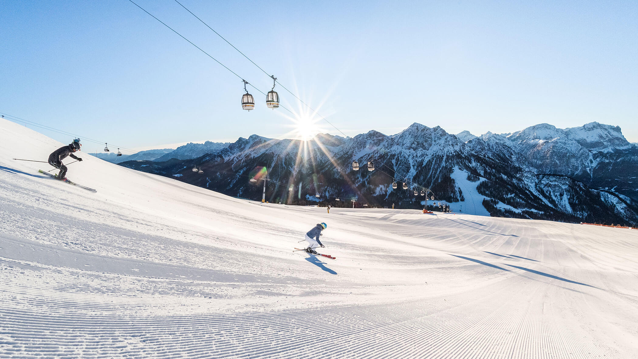 Two people skiing down a sunny, snowy mountain slope with chairlifts and mountain peaks in the background. - Kerschbaumerhof