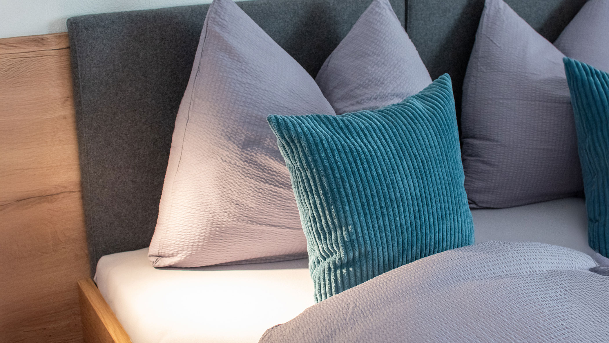 A neatly made bed with gray pillows and a textured teal cushion against a gray headboard. - Kerschbaumerhof