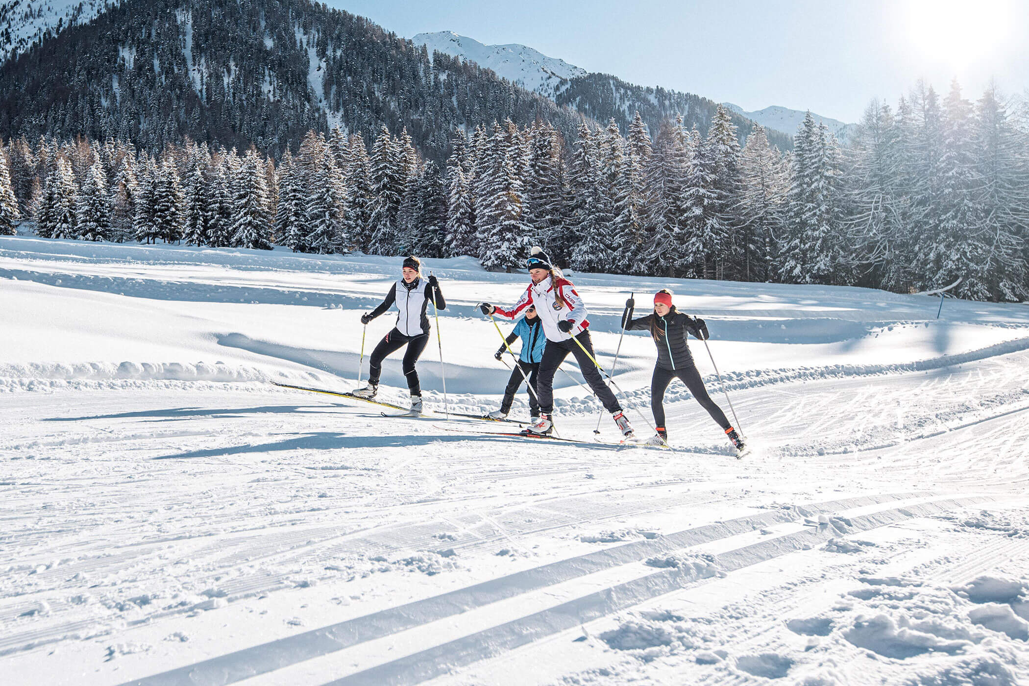 Vier Personen beim Skilanglauf auf einer verschneiten Loipe mit Bergen und Kiefern im Hintergrund. - Kerschbaumerhof