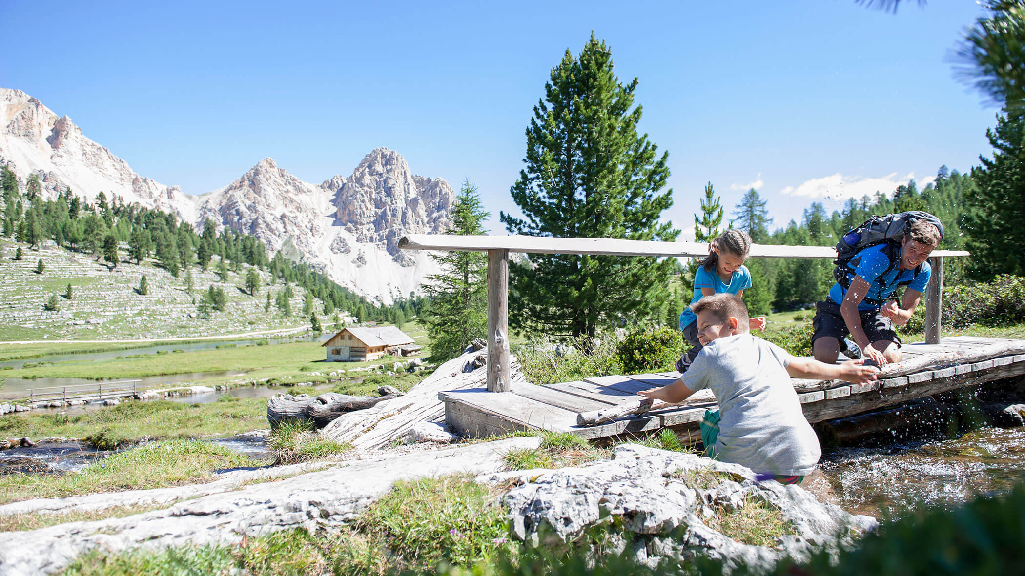 Eine Familie genießt die Natur auf einer kleinen Holzbrücke über einen Bach mit Bergen und Bäumen im Hintergrund. - Kerschbaumerhof