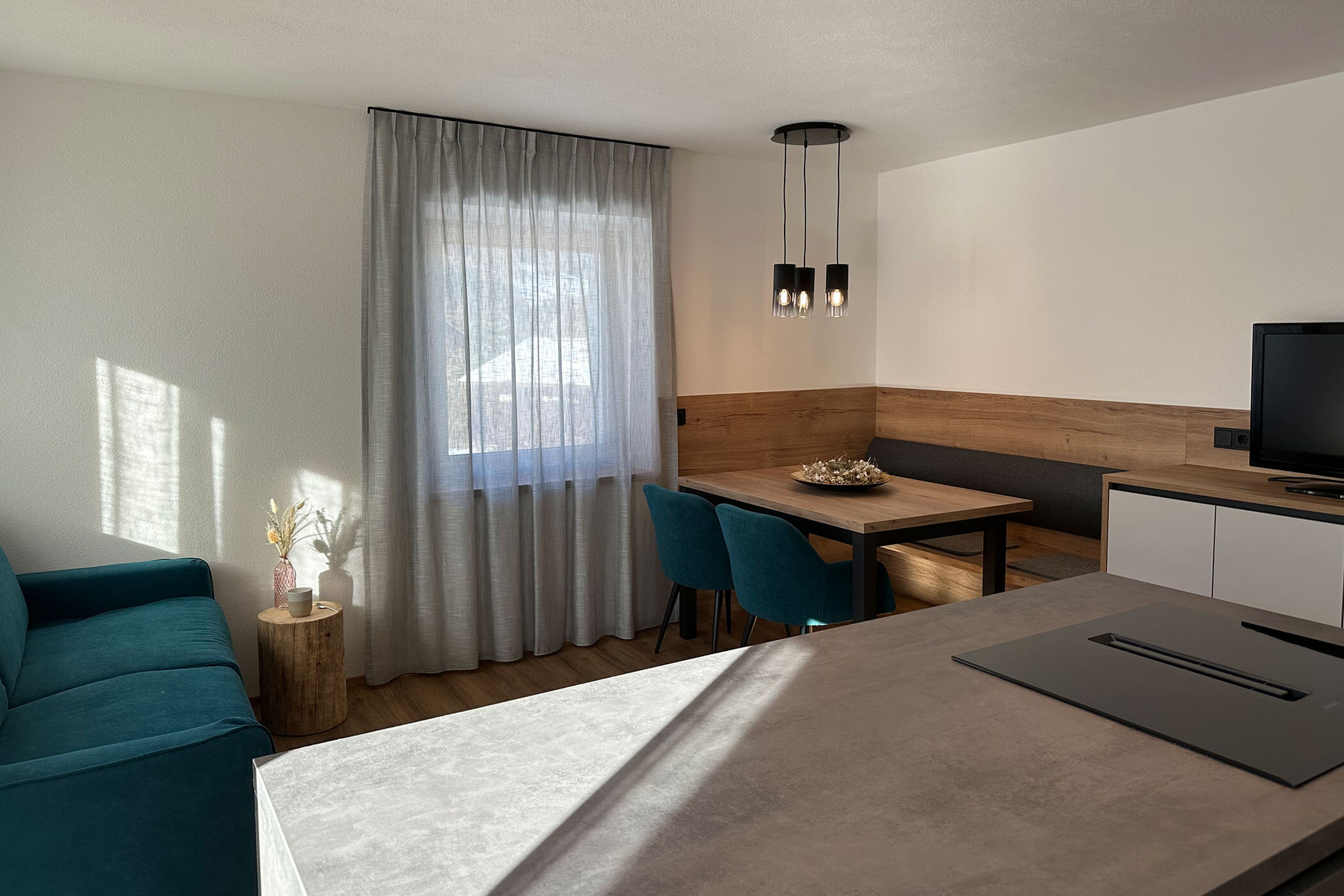 Moderner Wohn- und Essbereich mit tealfarbenen Stühlen, Holztisch und Sonnenlicht, das durch ein Fenster mit transparenten Vorhängen fällt. - Kerschbaumerhof