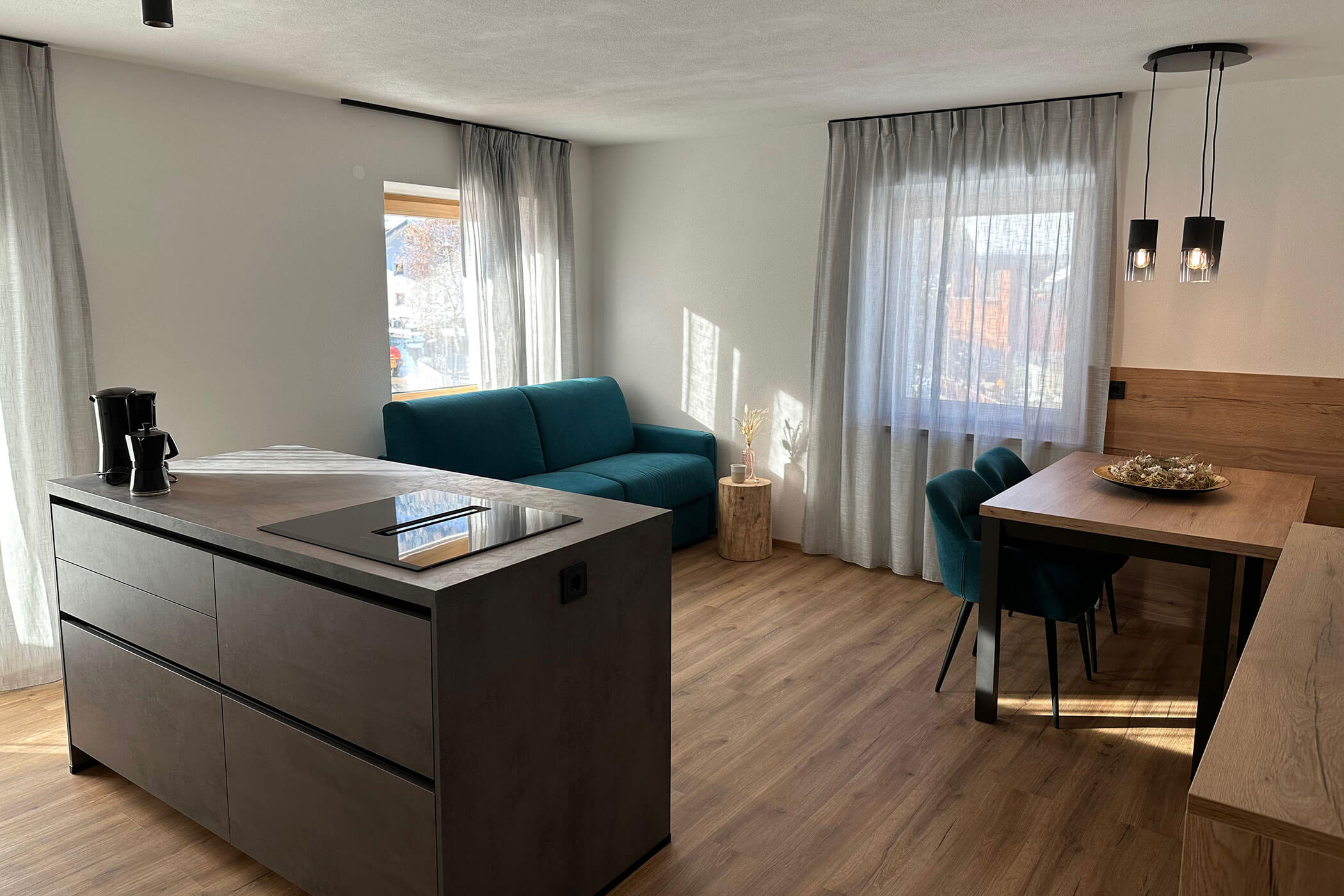 Modern, sunlit living and dining area with teal sofa, wood floors, and kitchen island. - Kerschbaumerhof