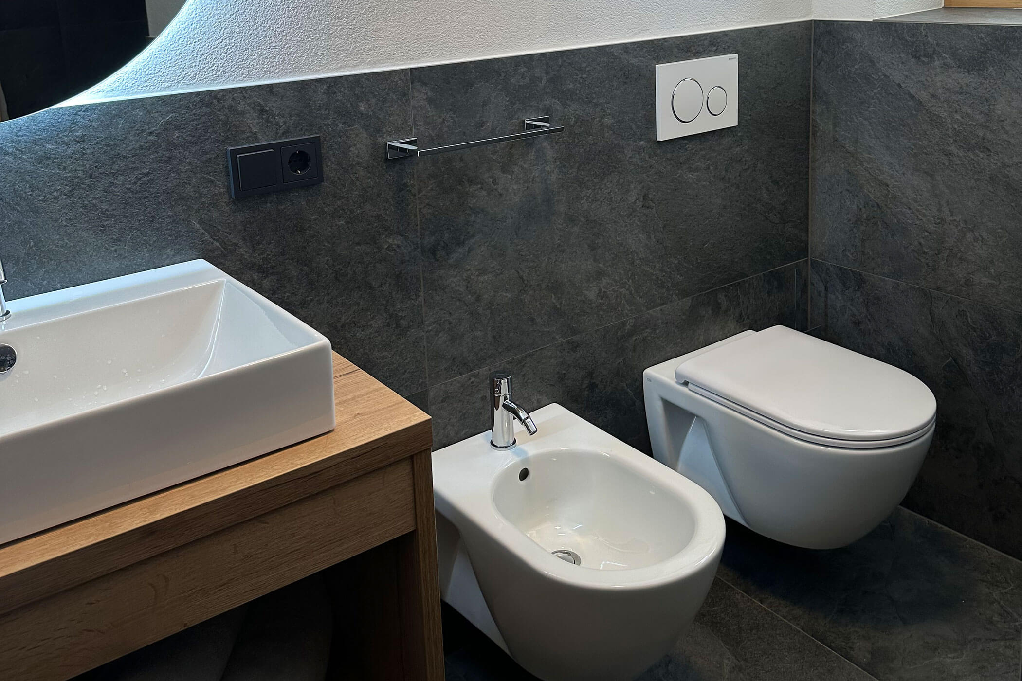 Modernes Badezimmer mit Waschbecken, Bidet und wandmontierter Toilette auf dunkelgrau gefliesten Wänden und Böden. - Kerschbaumerhof