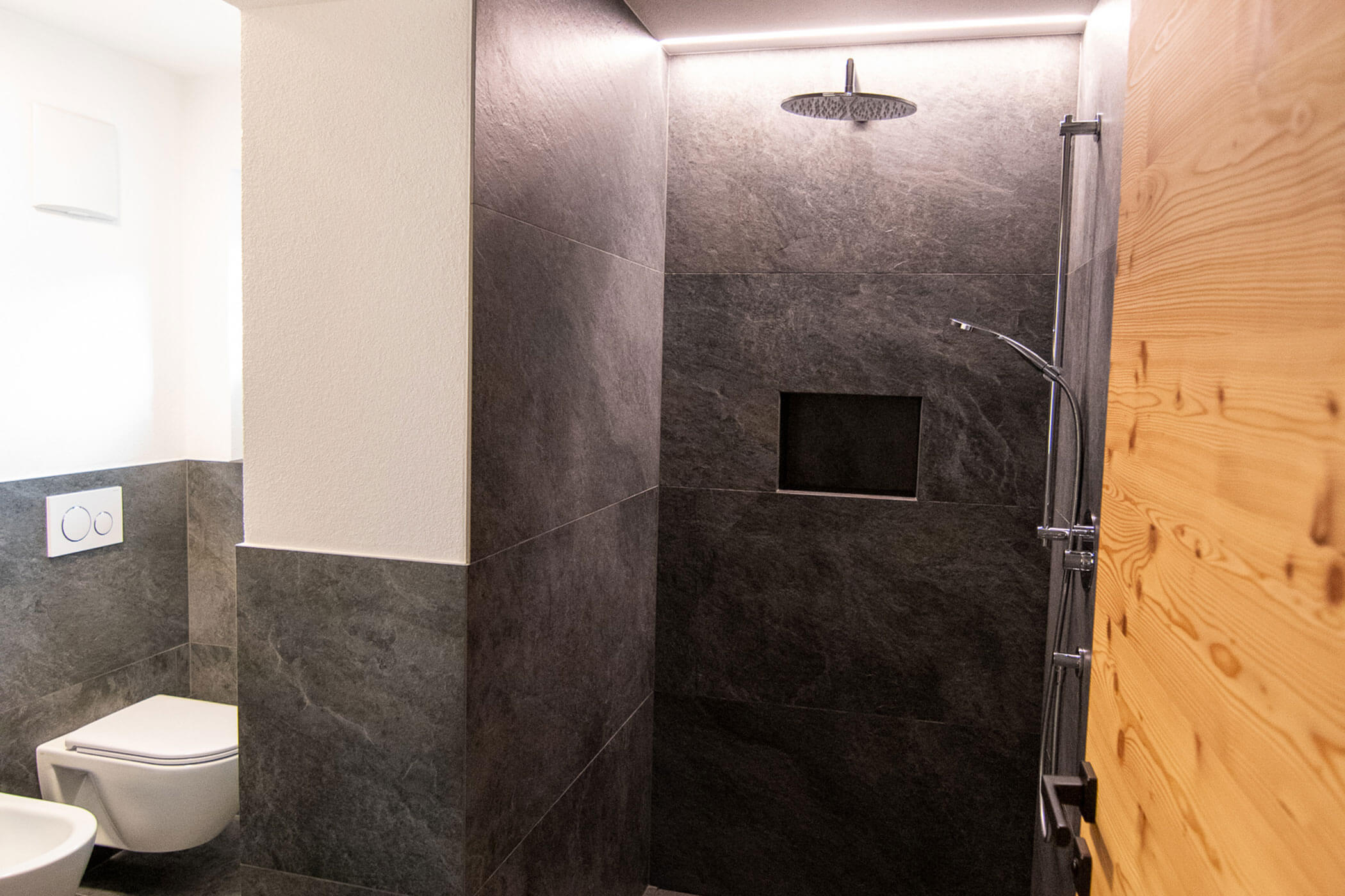 Modernes Badezimmer mit wandmontierter Toilette und begehbarer Dusche mit dunklen Fliesen und Regendusche. - Kerschbaumerhof