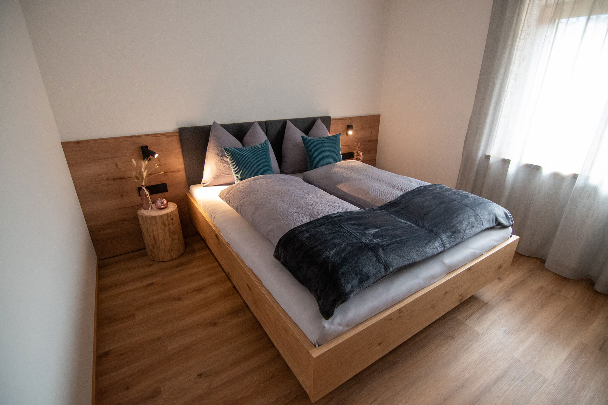 Modernes Schlafzimmer mit Holzboden, Doppelbett, grauer Bettwäsche, blauen Kissen und natürlichem Licht durch ein Fenster. - Kerschbaumerhof