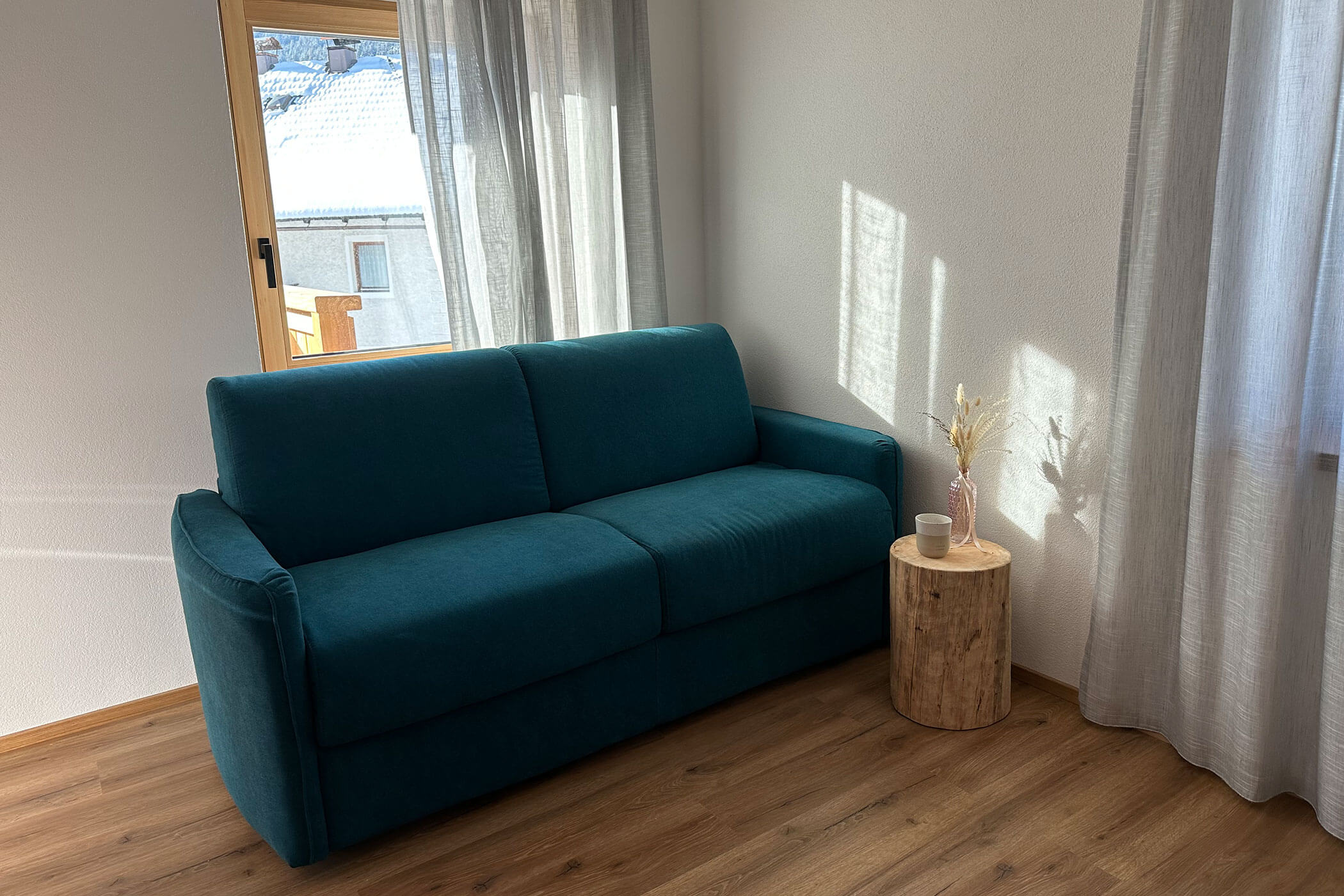 Ein tealfarbenes Sofa neben einem kleinen runden Holztisch mit Trockenblumen in einem hellen, sonnenbeschienenen Raum. - Kerschbaumerhof