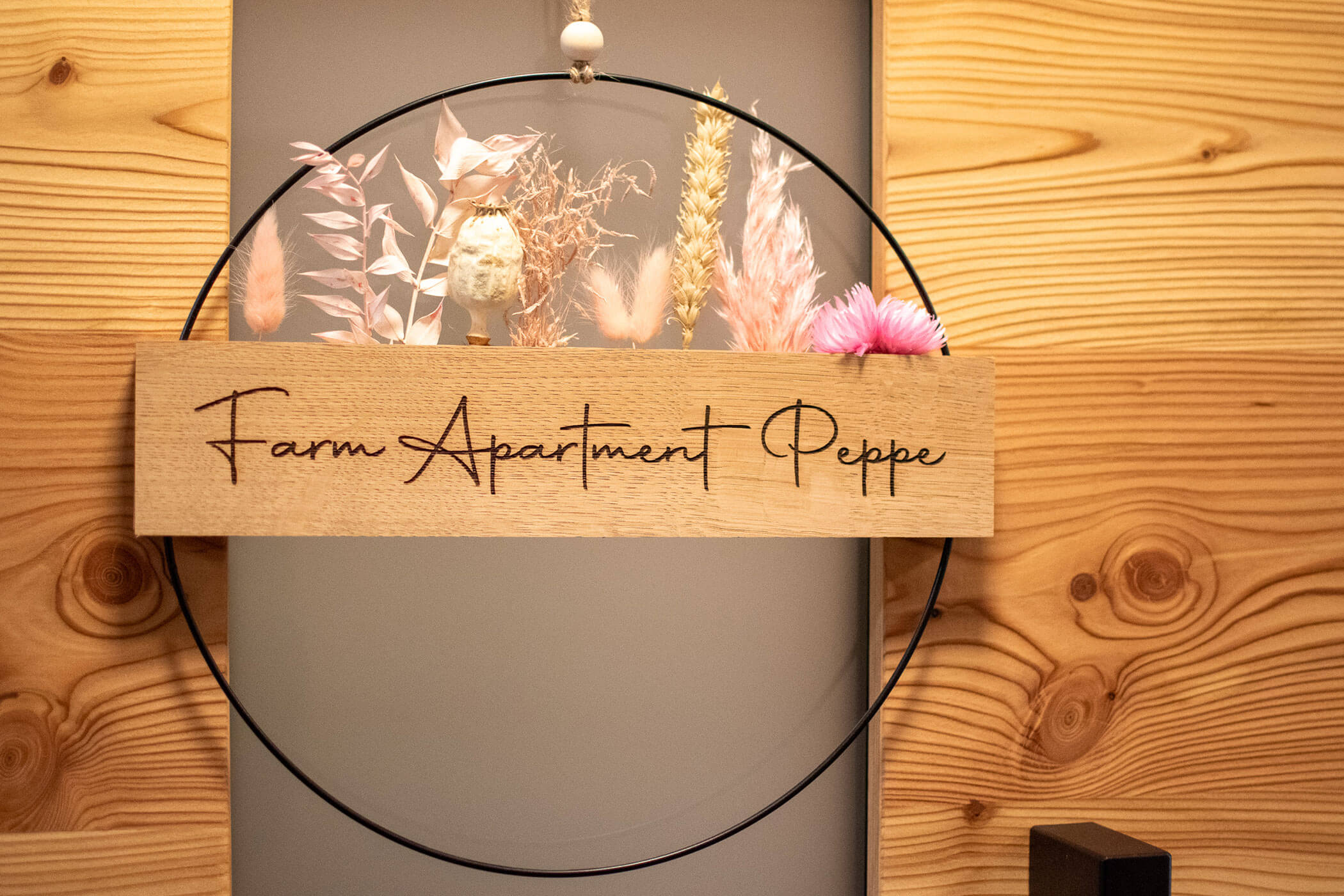 An einer Holzwand hängt ein mit Trockenblumen verziertes Holzschild mit der Aufschrift Bauernhof Peppe. - Kerschbaumerhof