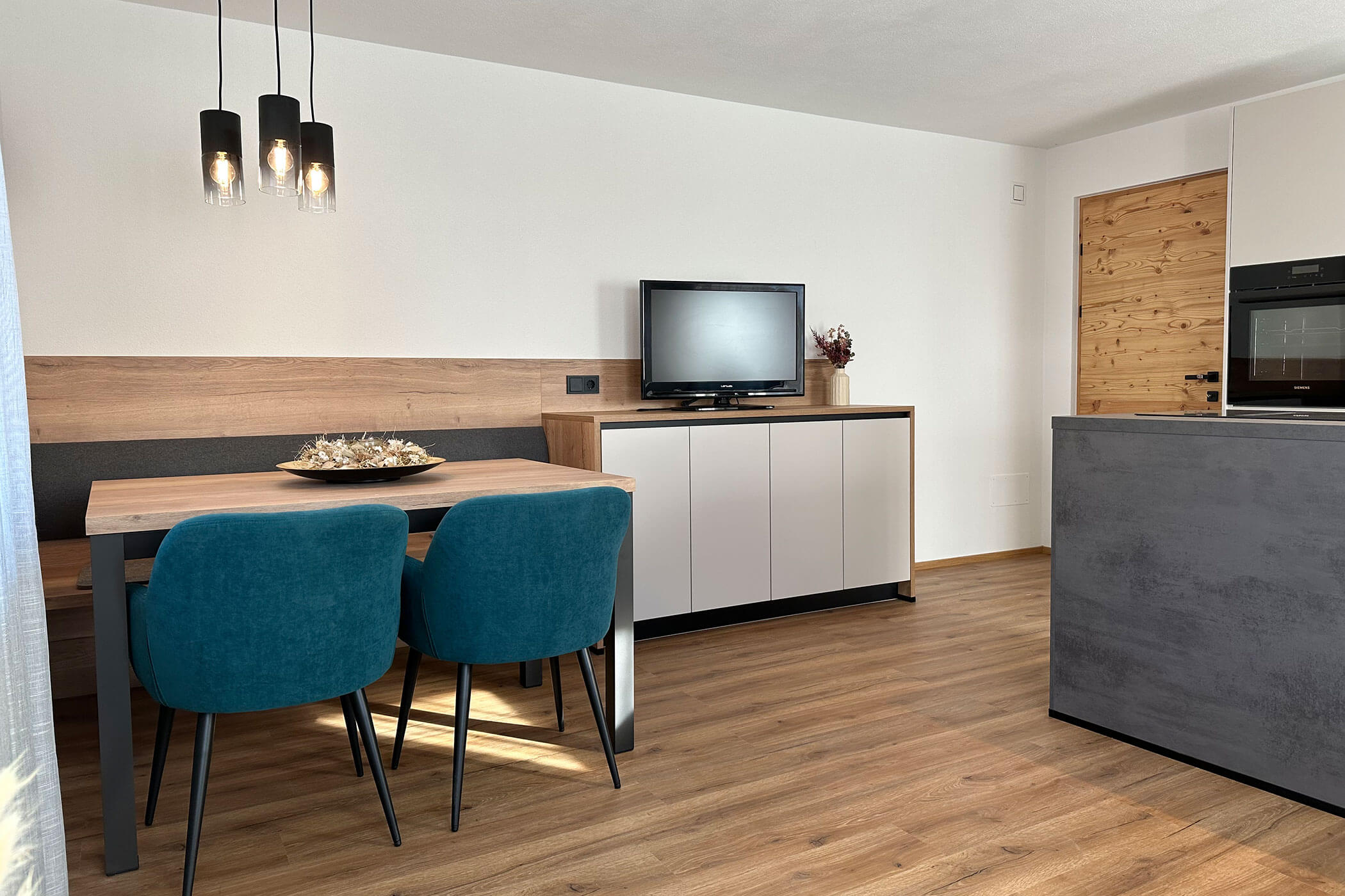 Moderner Essbereich mit tealfarbenen Stühlen, Holztisch, TV auf einem Sideboard und Pendelleuchten. - Kerschbaumerhof