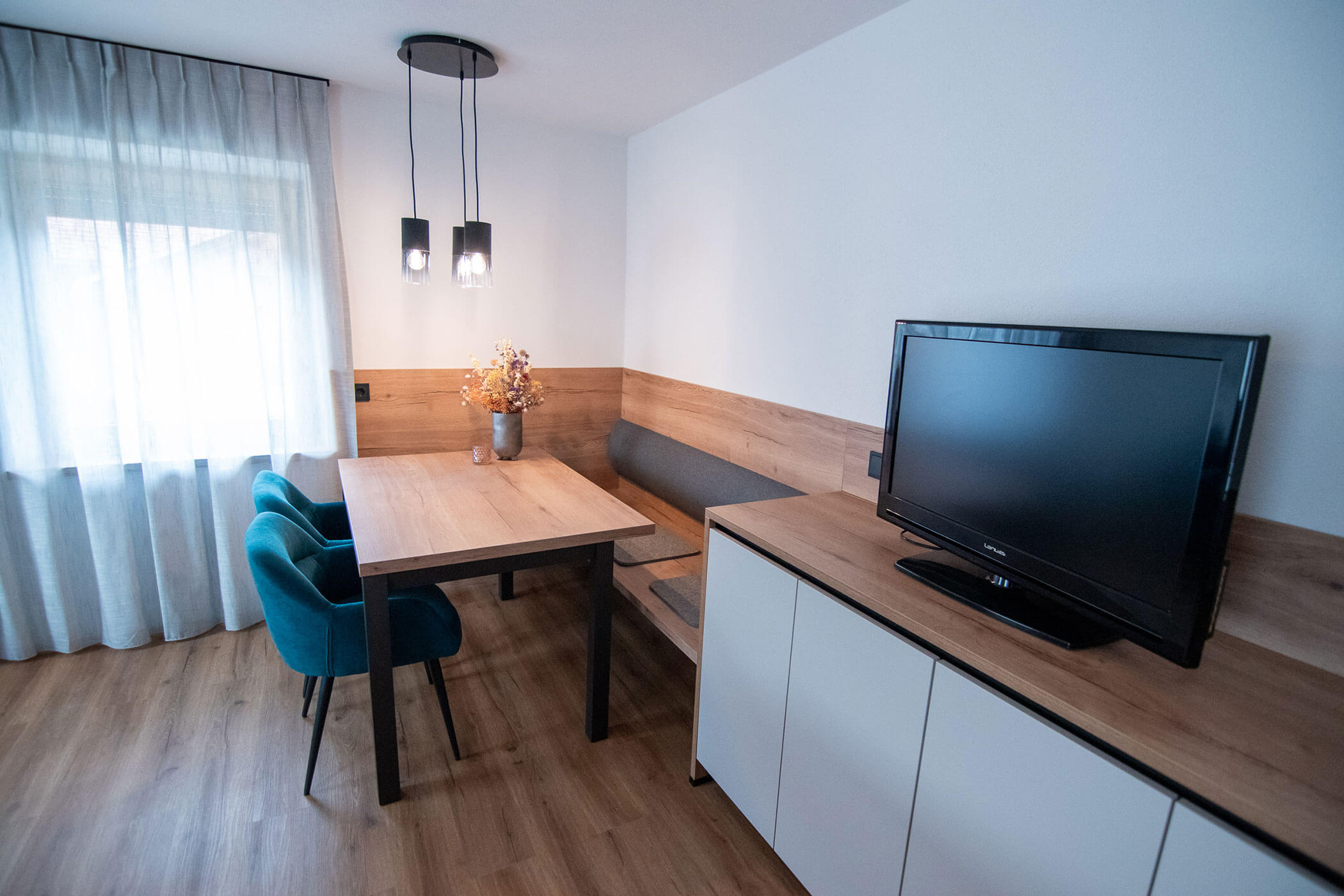 Moderne Essecke mit Holztisch, tealfarbenen Stühlen, Sitzbank, TV auf Schrank und Fenster mit transparenten Vorhängen. - Kerschbaumerhof