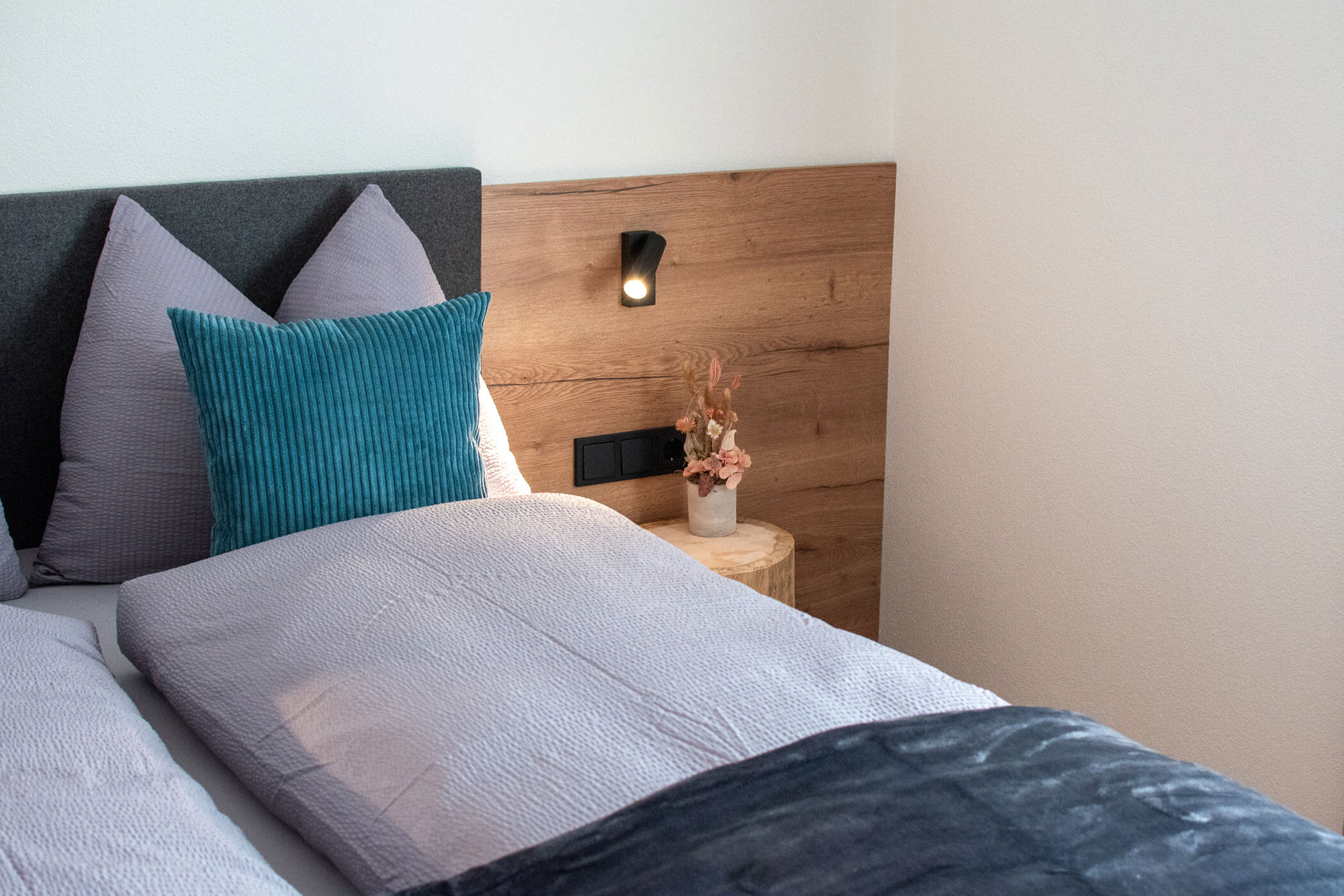 Ein ordentlich gemachtes Bett mit violettem Bettzeug, blauem Kissen, Kopfteil aus Holz, Leselampe und einer Vase mit Trockenblumen. - Kerschbaumerhof