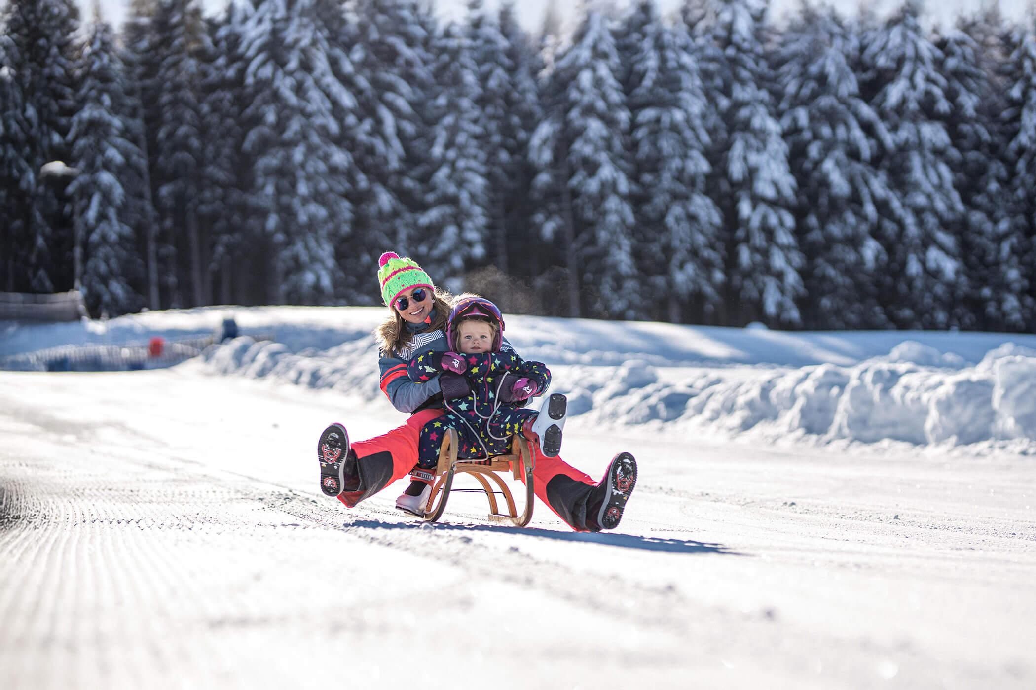 Eine Frau und ein Kind rodeln gemeinsam im Schnee mit einem verschneiten Wald im Hintergrund. - Kerschbaumerhof