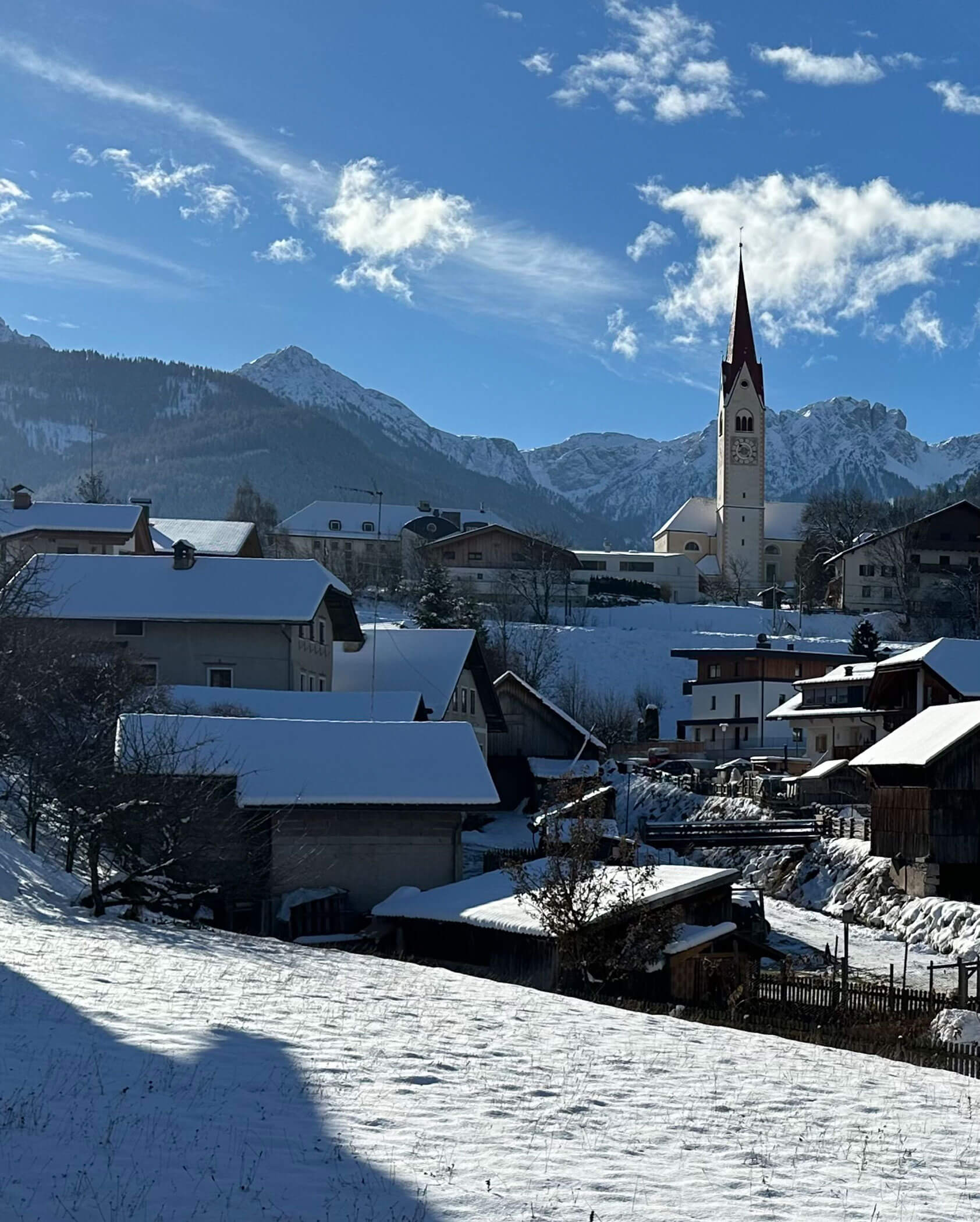 Schneebedecktes Dorf mit einem hohen Kirchturm, Bergen und blauem Himmel im Hintergrund. - Kerschbaumerhof