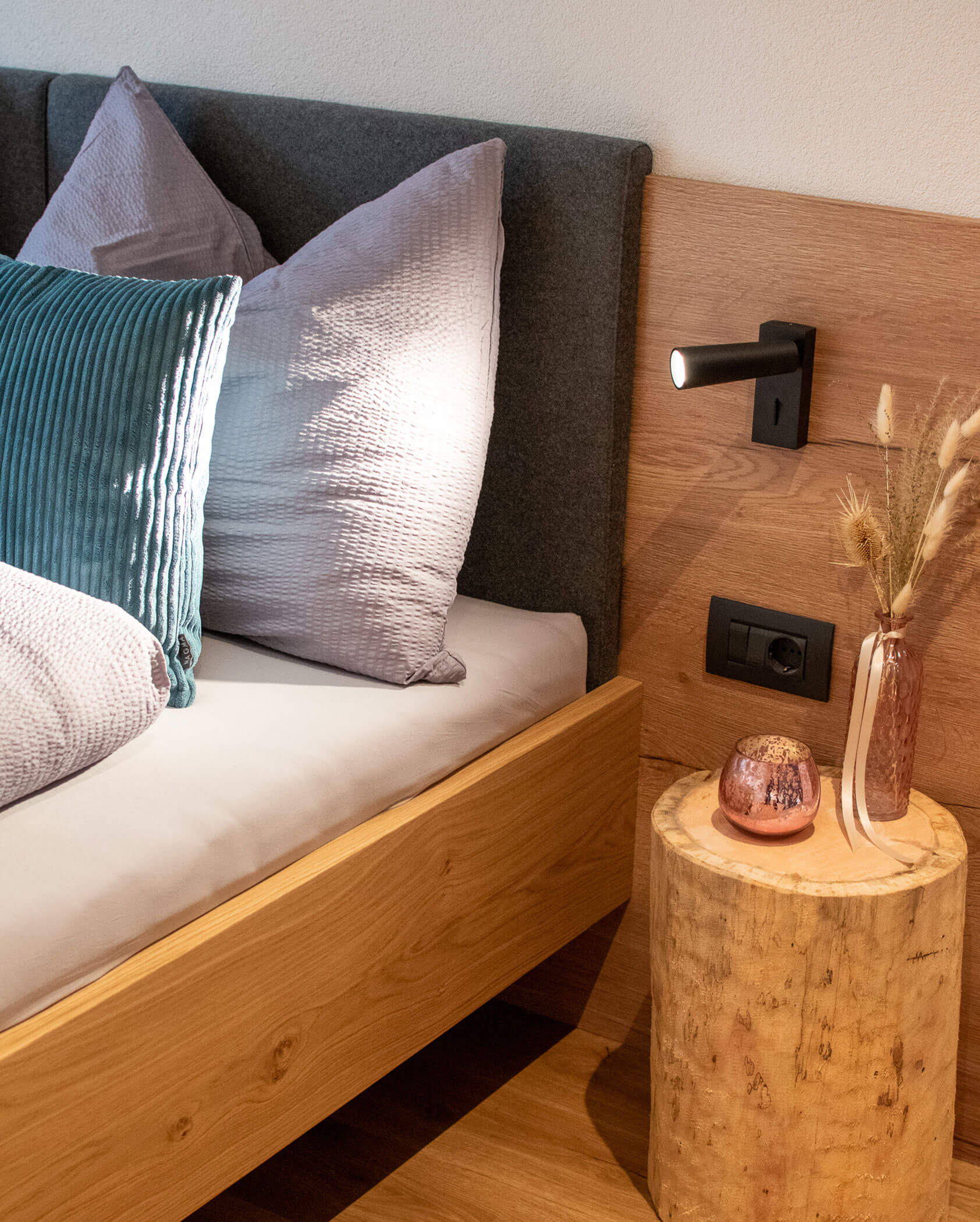 Bett mit Kissen, Nachttisch mit Holzstumpf, Trockenblumen in einer Vase und Wandleuchte in einem gemütlichen Schlafzimmer. - Kerschbaumerhof