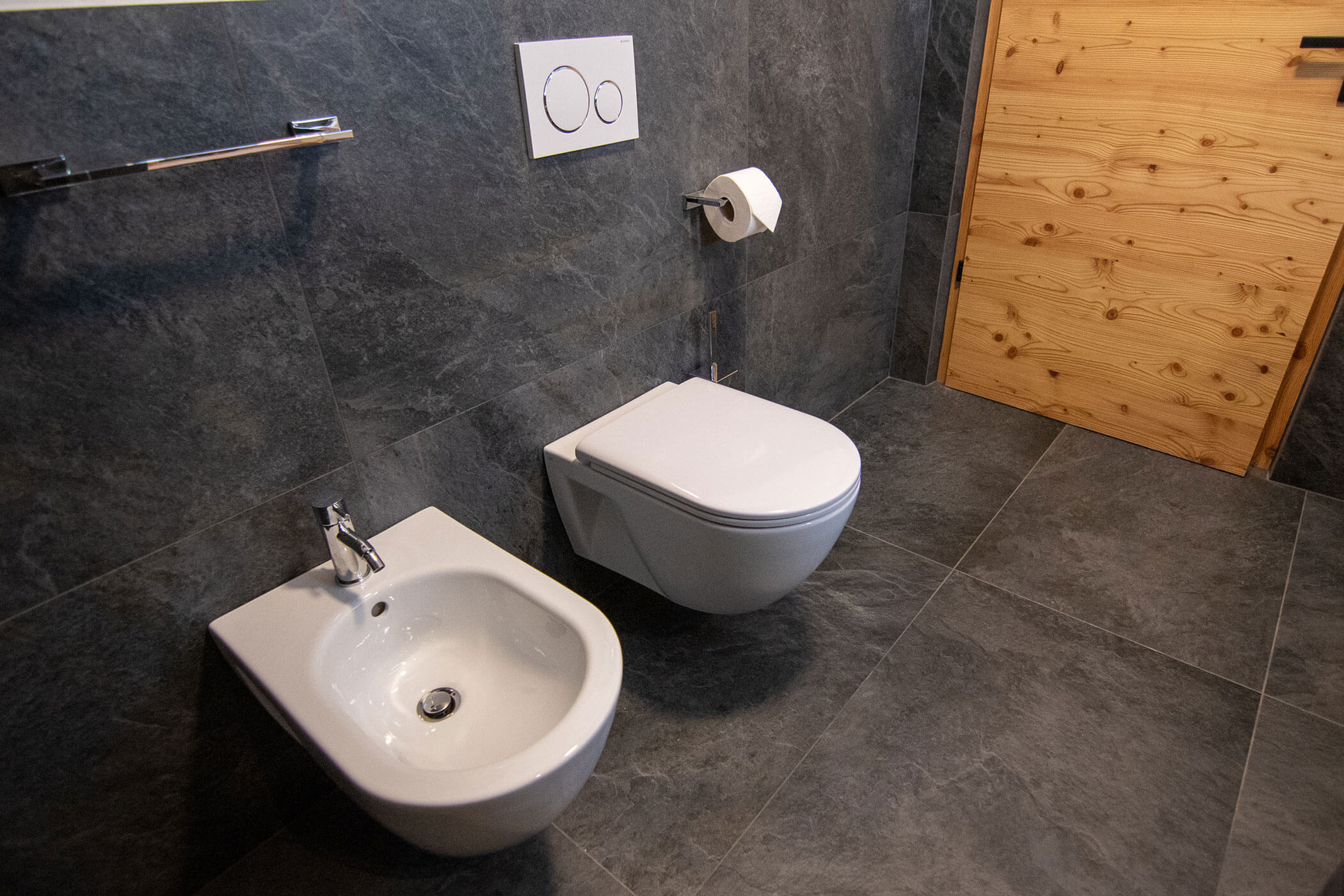 Modernes Badezimmer mit grauen Fliesen, einem Bidet, einer Wandtoilette und einer Holztür. - Kerschbaumerhof