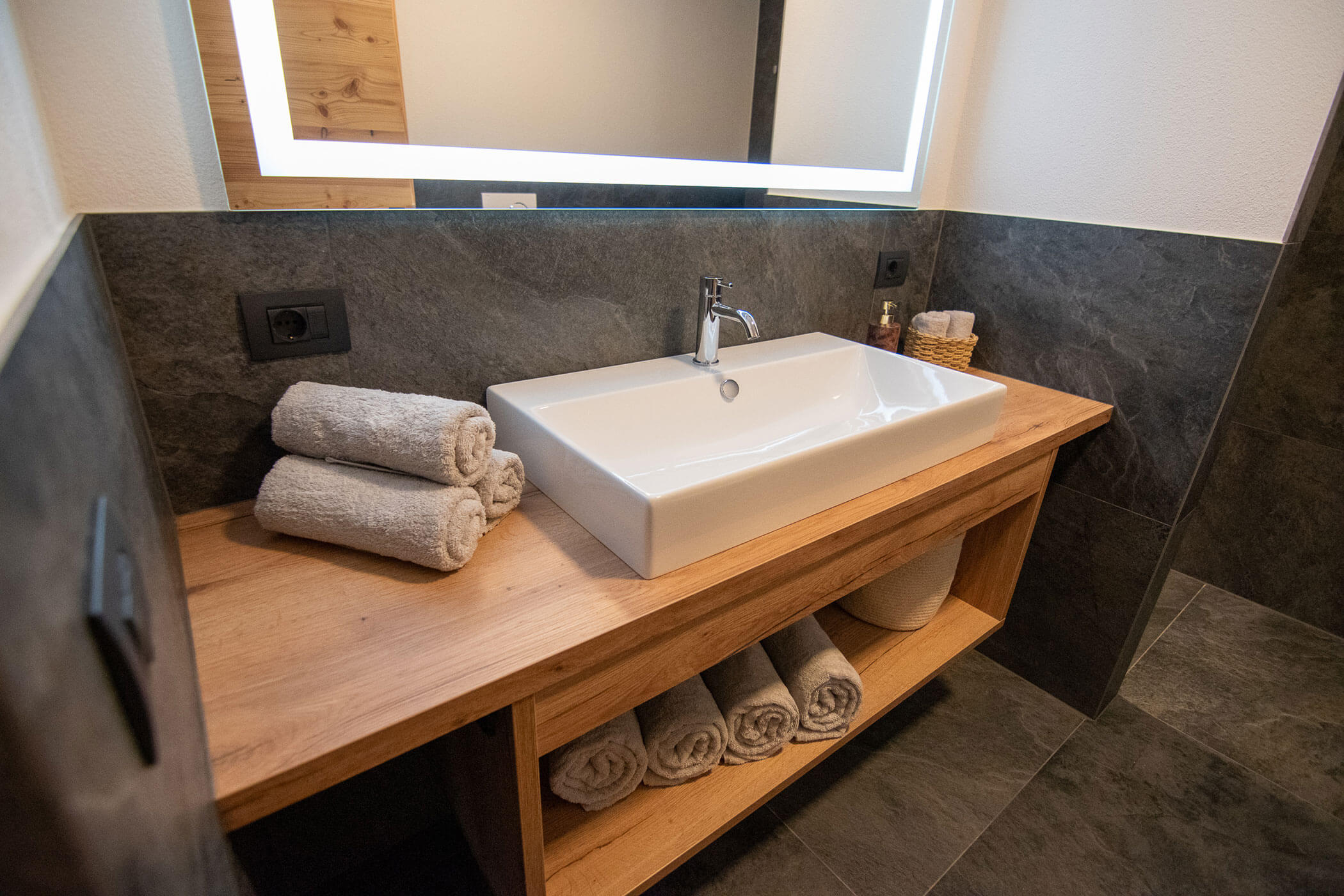 Modernes Badezimmer mit rechteckigem Waschbecken, gerollten Handtüchern und Holzarbeitsplatte auf Schieferfliesenboden. - Kerschbaumerhof