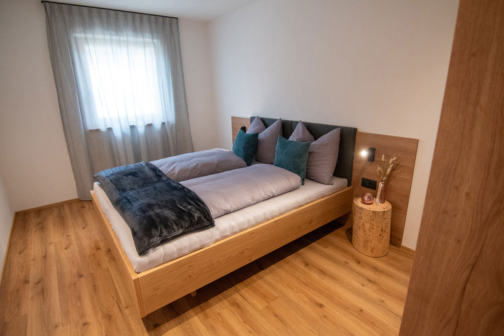 Minimalistisches Schlafzimmer mit einem Holzbett, grauer Bettwäsche, Kissen, einem kleinen Beistelltisch und durchsichtigen Vorhängen am Fenster. - Kerschbaumerhof