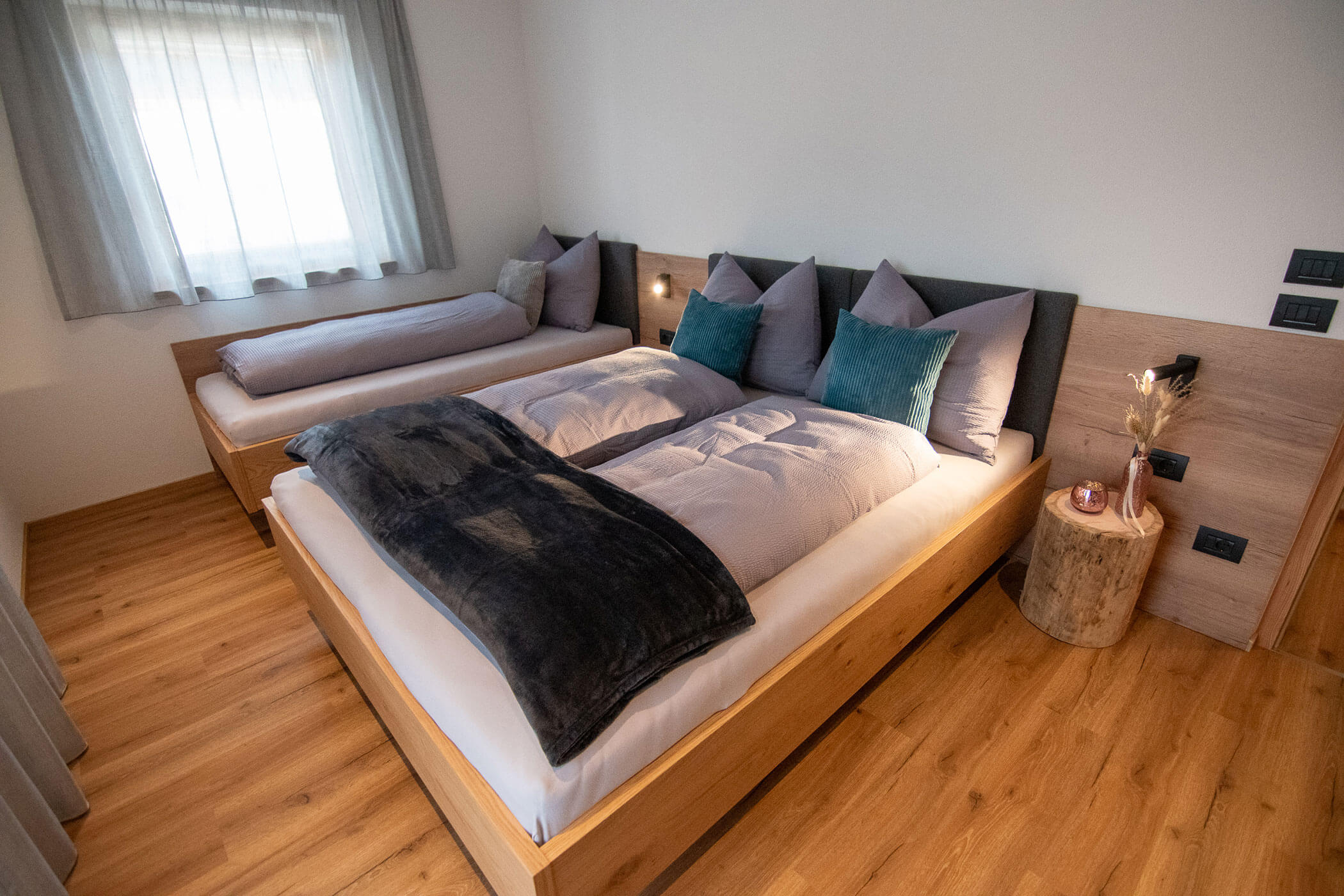 Modernes Schlafzimmer mit Holzboden, zwei Betten, grauer Bettwäsche und einem Nachttisch aus einem Baumstumpf. - Kerschbaumerhof