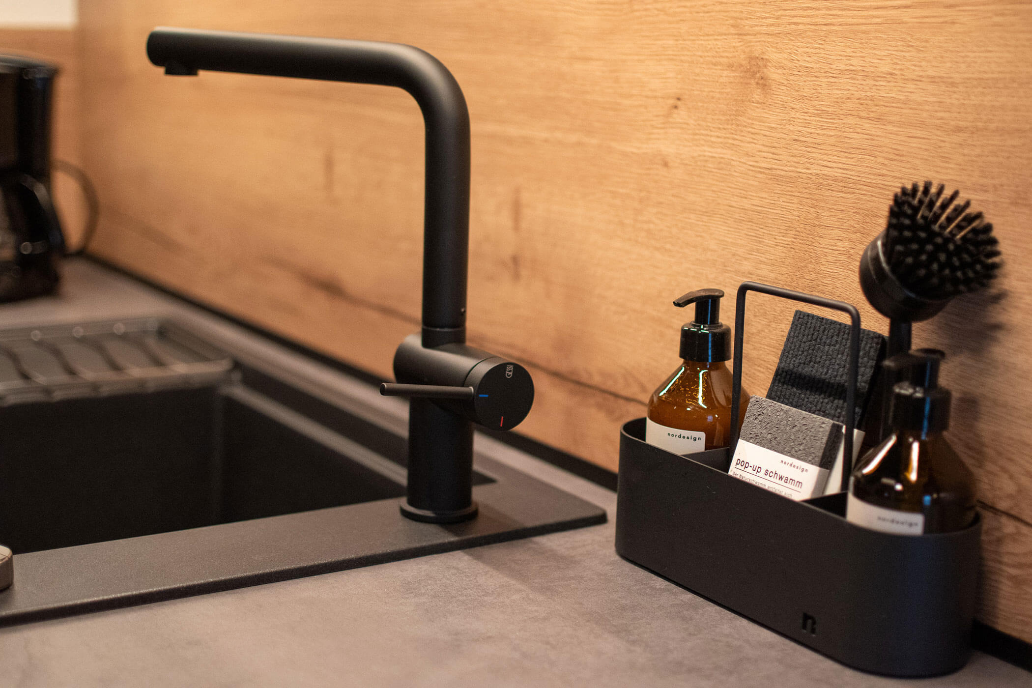 Schwarzer Küchenhahn neben einem Waschbecken, mit Seife, Bürste und Handtüchern in einem Tablett auf dem Tresen vor einer hölzernen Aufkantung. - Kerschbaumerhof