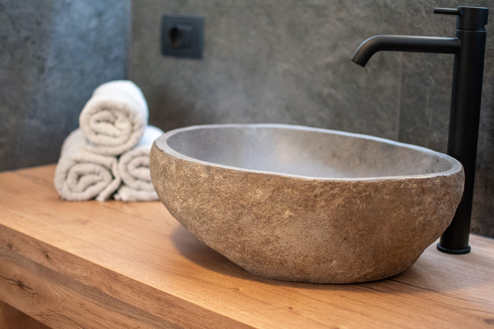 Waschbecken aus Stein mit schwarzem Wasserhahn auf einem hölzernen Tresen, daneben drei gerollte weiße Handtücher. - Kerschbaumerhof