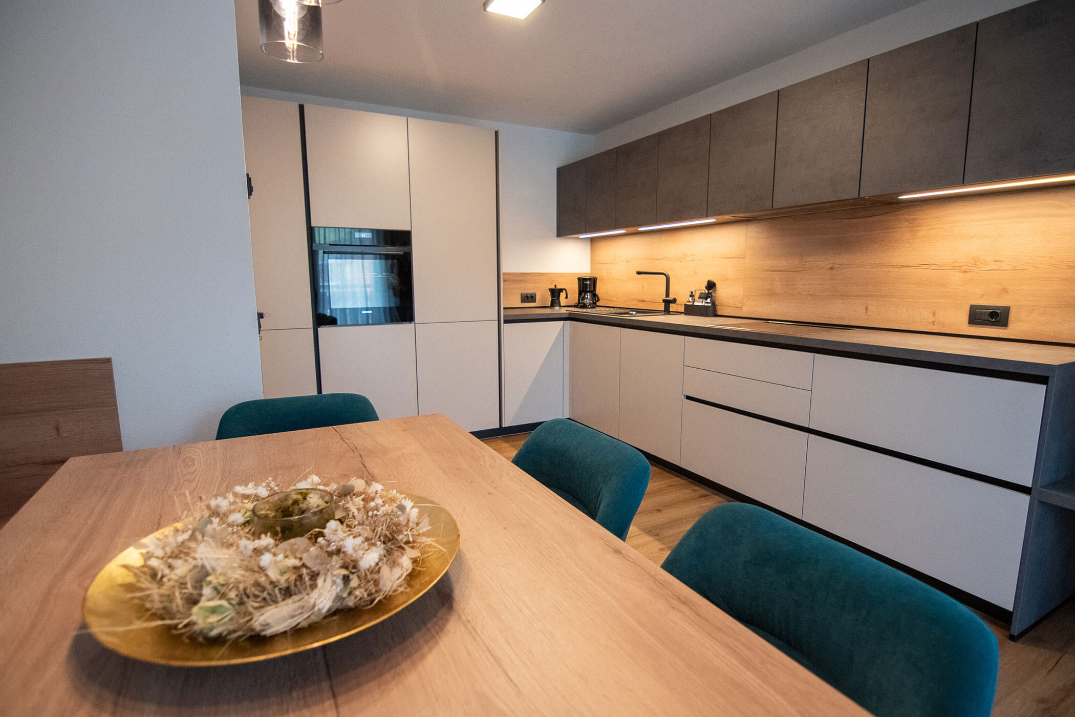 Moderne Küche mit weißen Schränken, Holzakzenten, Stühlen in Petrol und einem dekorativen Mittelstück auf dem Esstisch. - Kerschbaumerhof