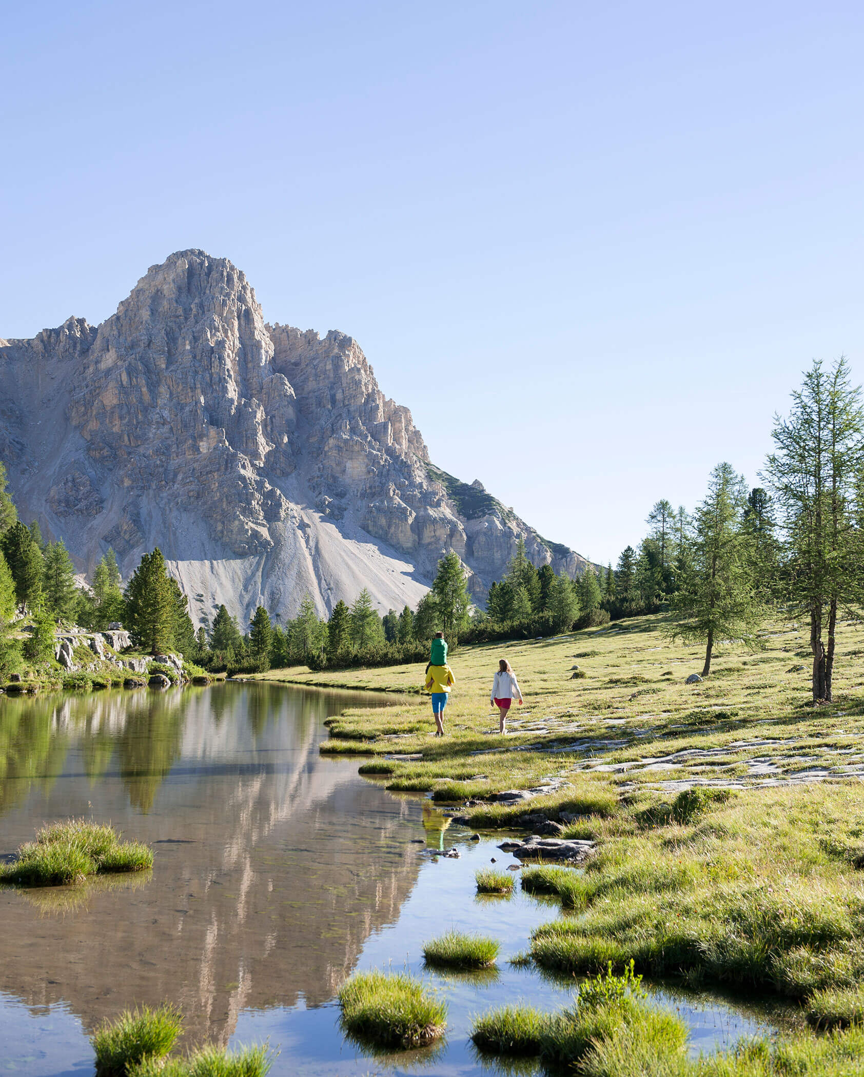 Zwei Personen spazieren an einem ruhigen See mit einem Berg und Bäumen im Hintergrund unter einem klaren Himmel. - Kerschbaumerhof